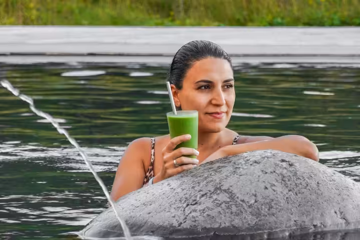 Woman relaxing in geothermal Vok baths holding green smoothie by stone edge in Iceland