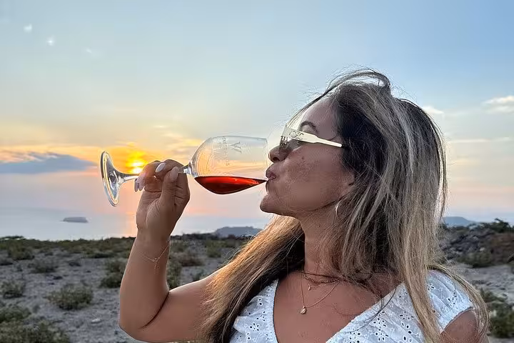 Woman sipping Santorini red wine at golden-hour caldera viewpoint on sunset winery tour with tapas pairing