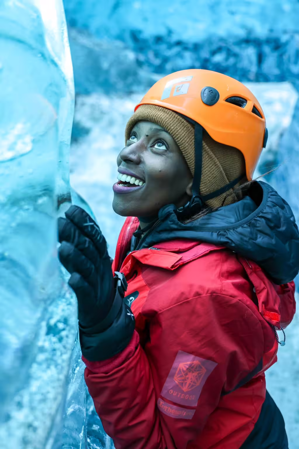 Smiling woman in red jacket and orange helmet touches vivid blue glacier ice wall on an ice cave tour