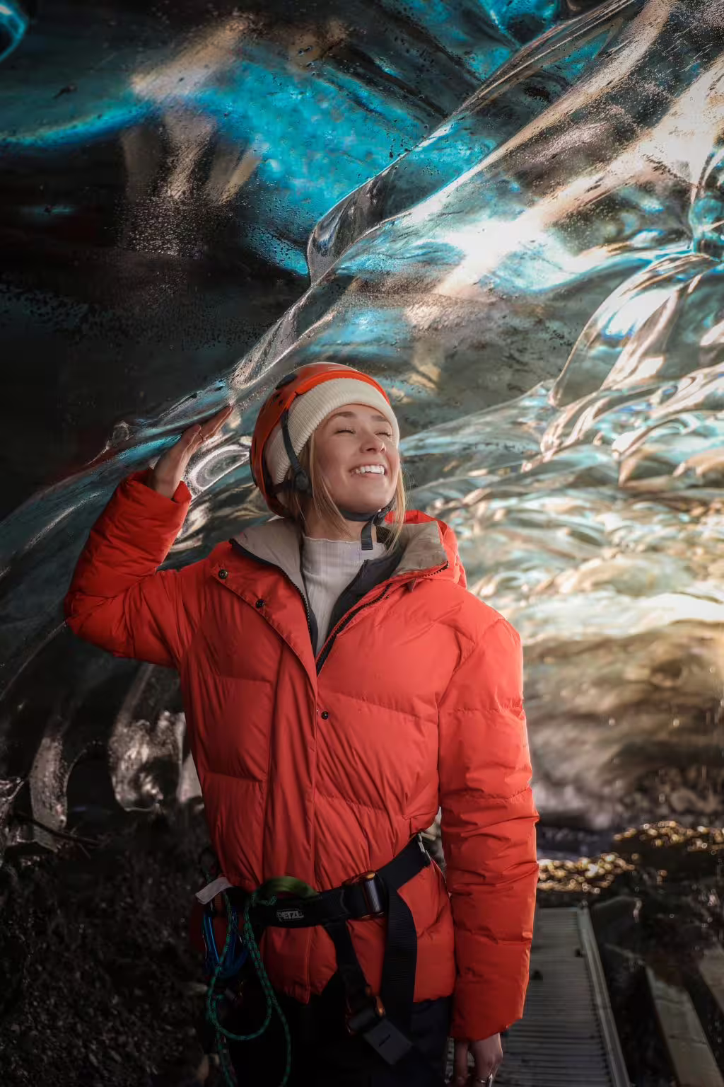A happy woman in a red jacket and safety helmet touches the crystal-blue glacial ice wall inside an Iceland sapphire ice cave guided tour.