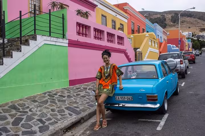 Woman posing with a vintage blue car in Bo-Kaap, surrounded by colorful, vibrant houses on a lively street.