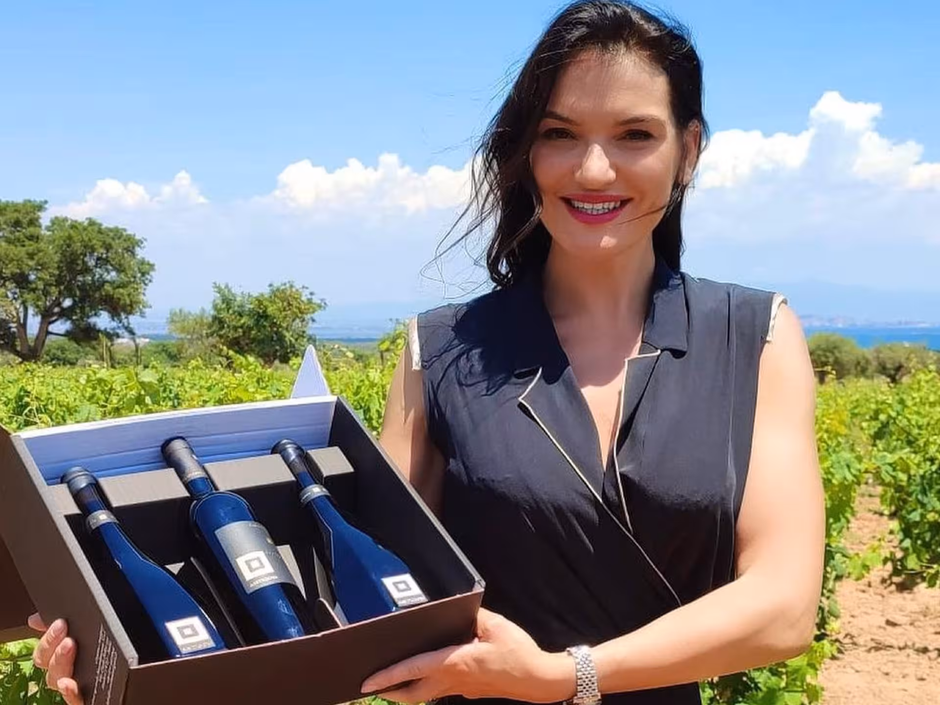 Smiling woman holding a wine gift set amidst scenic vineyards in Cagliari countryside hills.