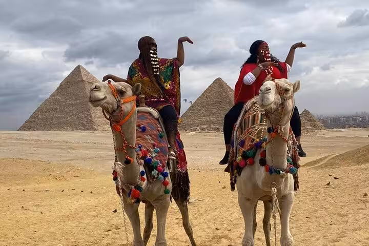 Women riding decorated camels in front of the Giza Pyramids on an all inclusive tour with pyramid entry and Sphinx visit