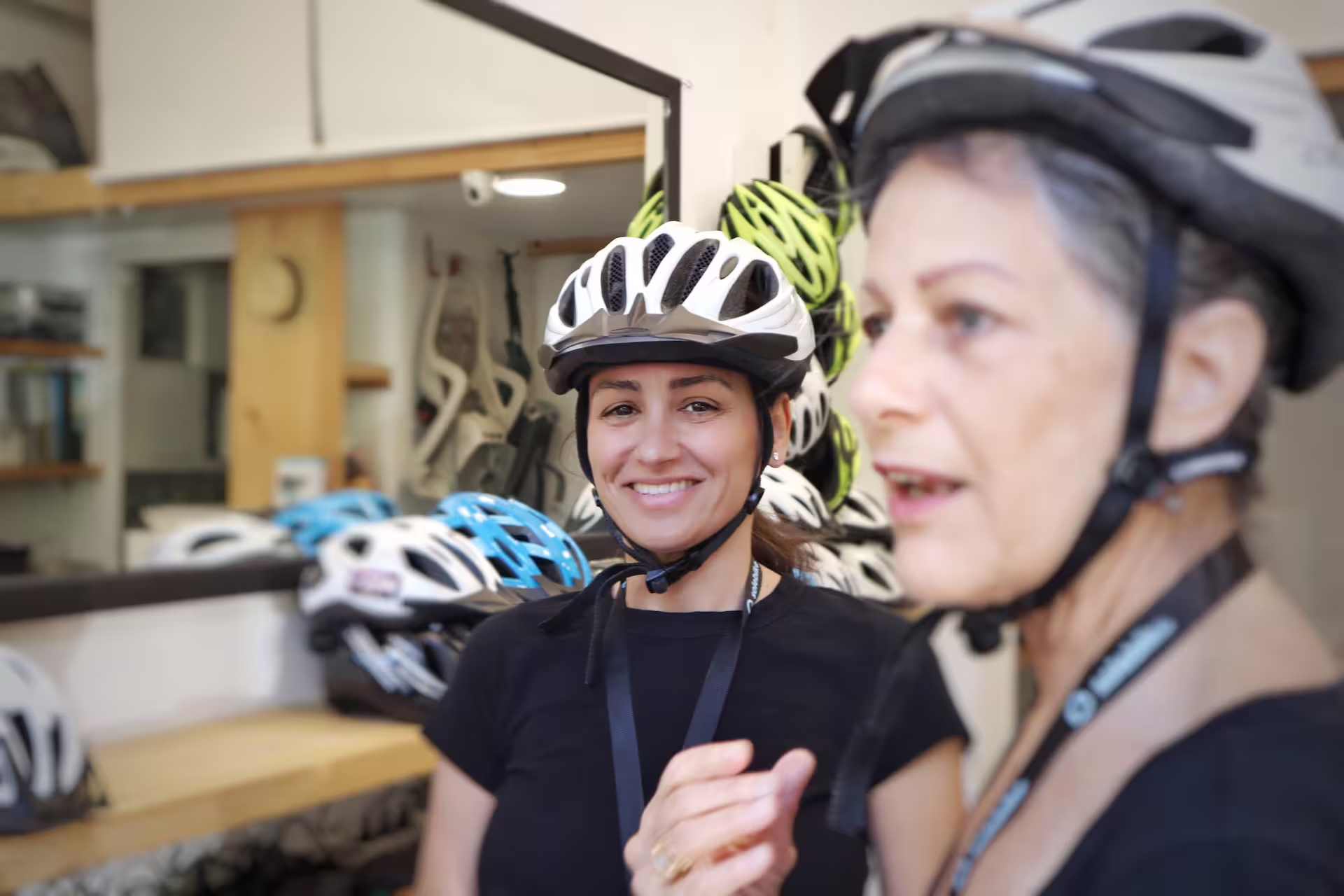 Two women wearing helmets smile inside a bike rental shop, preparing for a guided e-bike tour in Athens.