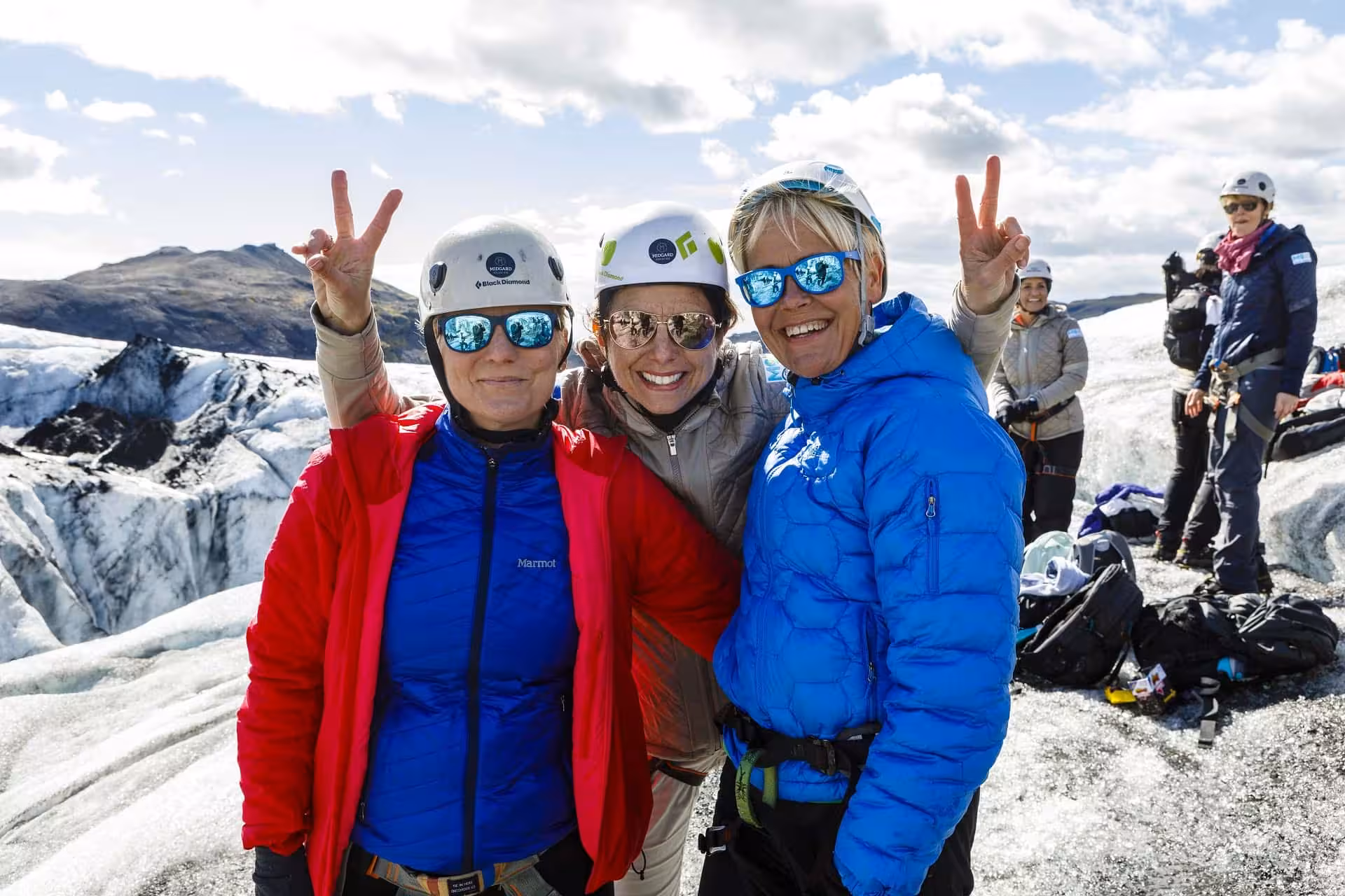 Three smiling women in helmets and sunglasses making peace signs on an Icelandic glacier