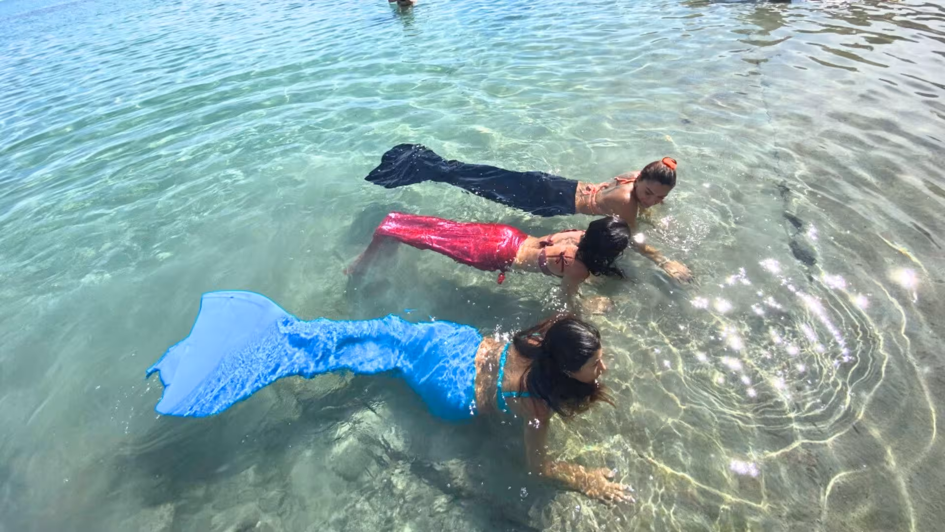 Three women in colorful mermaid tails enjoy swimming in Cagliari's clear Gulf of Angels waters.