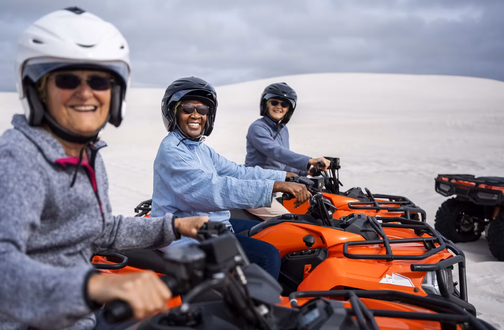 Three women wearing helmets riding orange ATV quad bikes across white sand dunes