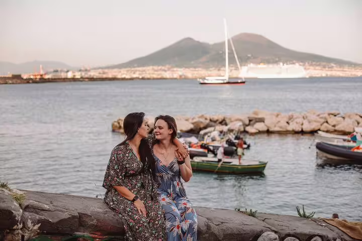 Two women sharing a joyful moment by the sea in Naples with Mount Vesuvius in the background during a private photoshoot.