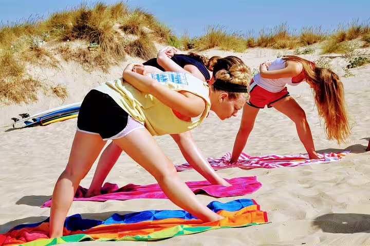 Women practicing yoga on sandy Caparica beach with colorful towels, enjoying a sunny surf and wellness retreat experience.
