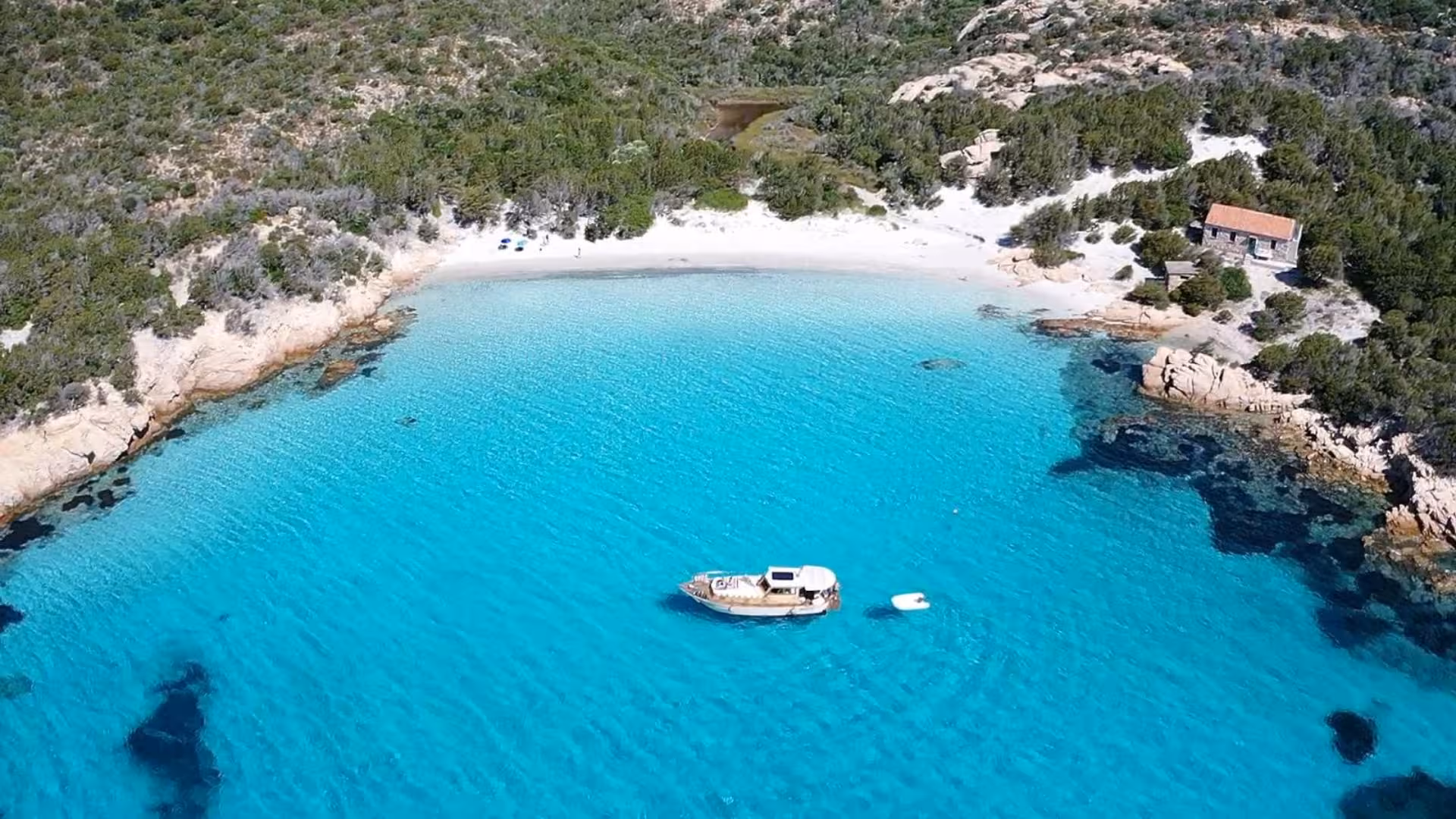 Aerial view of a wooden boat in crystal-clear blue waters near a secluded beach in the La Maddalena archipelago.