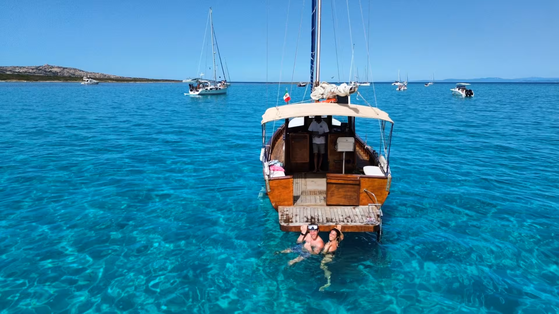 Tourists enjoying a swim beside a wooden boat in the crystal-clear waters of Stintino near Asinara Island.