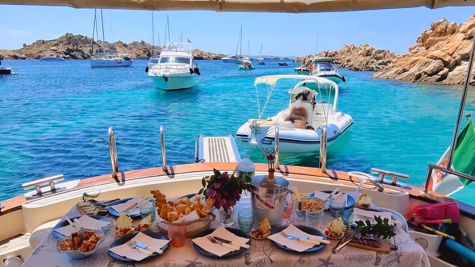 Gourmet dining setup on a wooden boat surrounded by turquoise waters and yachts in La Maddalena, Corsica.