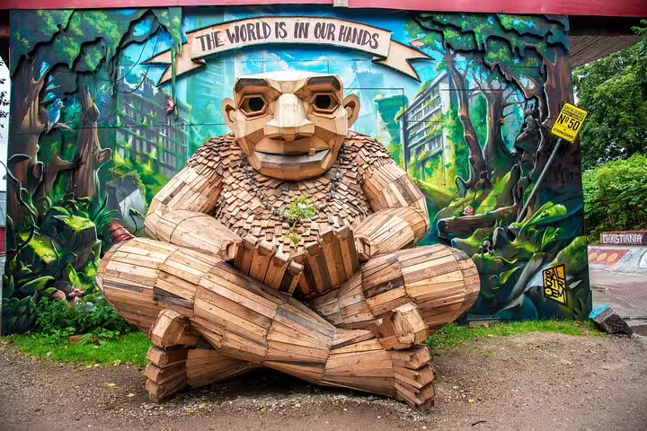 Large wooden troll sculpture sitting cross-legged in front of colorful mural in Christiania Copenhagen