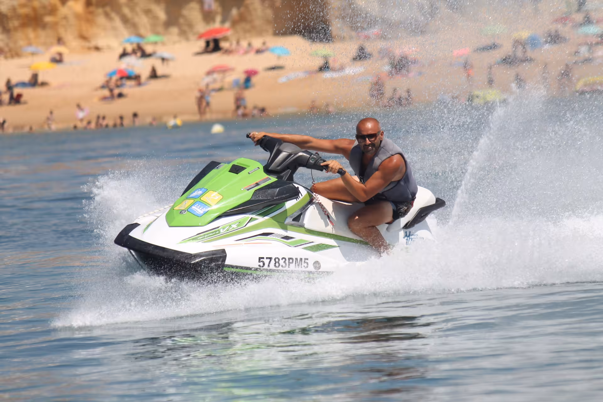 Man riding a green and white Yamaha jet ski at high speed near a crowded sandy beach on a sunny day