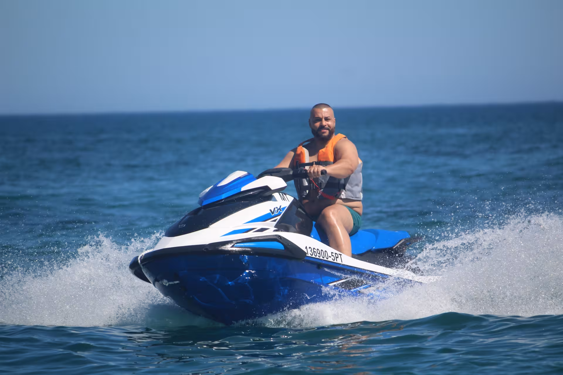 Man wearing an orange life vest riding a blue and white Yamaha jet ski across open ocean on a clear day