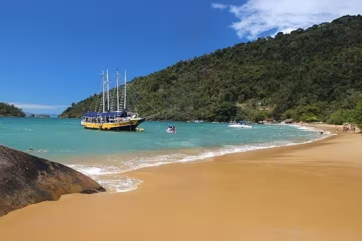 Scenic view of a yellow boat anchored near a lush green island with sandy beach on Paraty Tours boat tour with lunch.