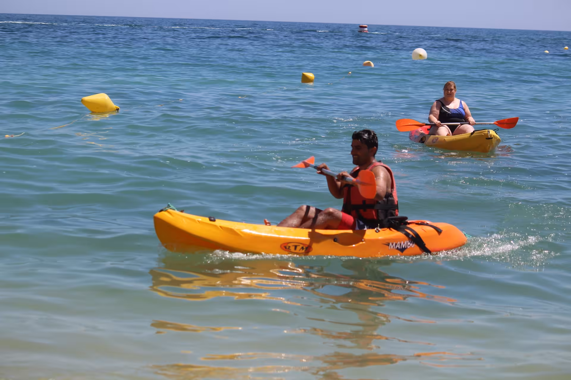 Two kayakers paddling yellow kayaks in calm turquoise ocean waters wearing life jackets on a sunny day