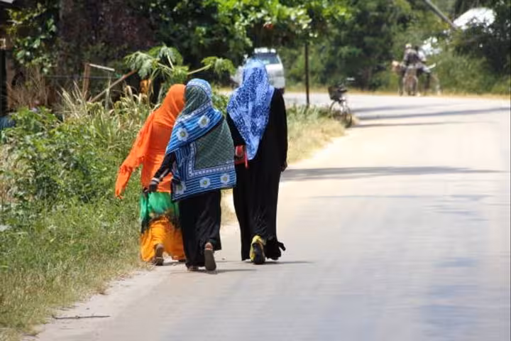 Three women in colorful hijabs walk along a scenic road in Zanzibar, showcasing local culture and vibrant attire.