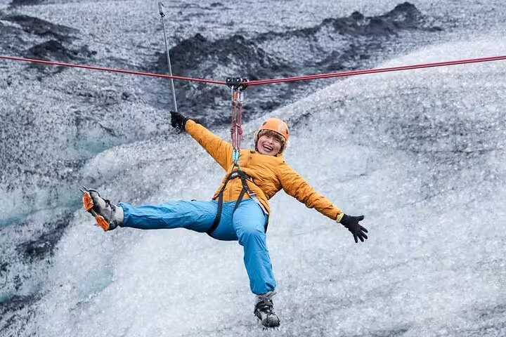 Excited adventurer ziplining over Iceland glacier lagoon wearing orange helmet and crampons on thrilling glacier tour