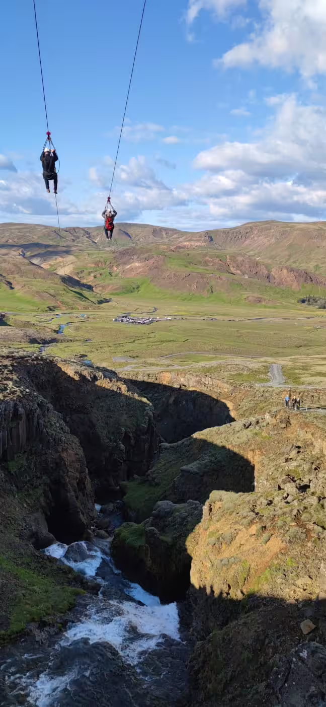 Two people ziplining above a scenic canyon with lush greenery and a flowing river under a clear blue sky.