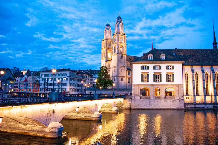 Grossmünster church twin towers illuminated at dusk over Limmat River with stone bridge in Zurich