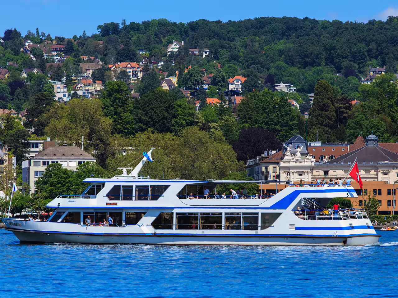 A passenger boat sailing on Lake Zurich with lush greenery and charming Swiss architecture visible in the background.