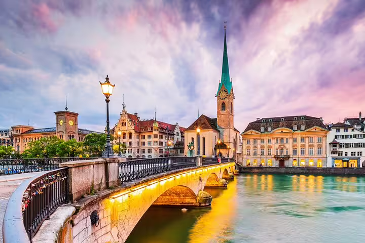Historic Zurich waterfront with Fraumünster church spire and illuminated stone bridge over Limmat River at dusk