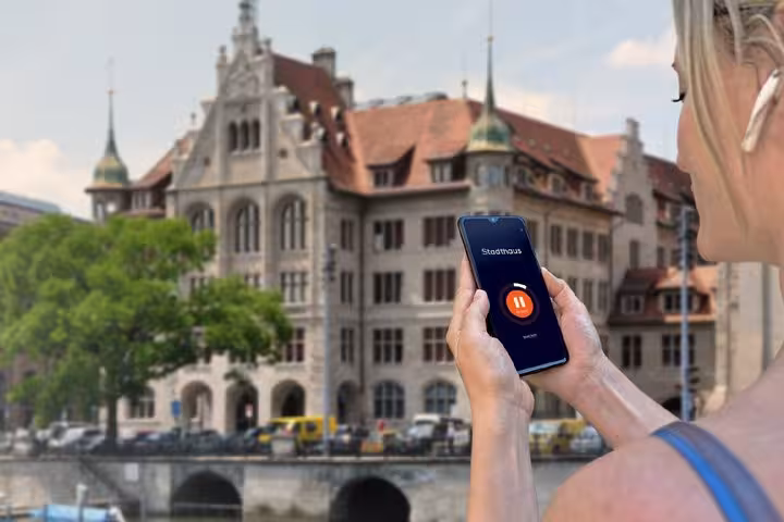 Woman holding smartphone with audio guide app in front of Zurich Stadthaus building by river