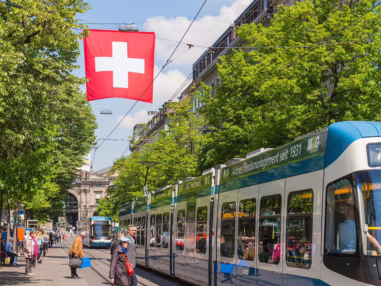 Swiss flag above a bustling Zurich street with trams and pedestrians on a sunny day during the private tour.